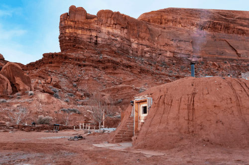 A small earthen structure with a wooden door sits against a backdrop of red rock formations and a blue sky.